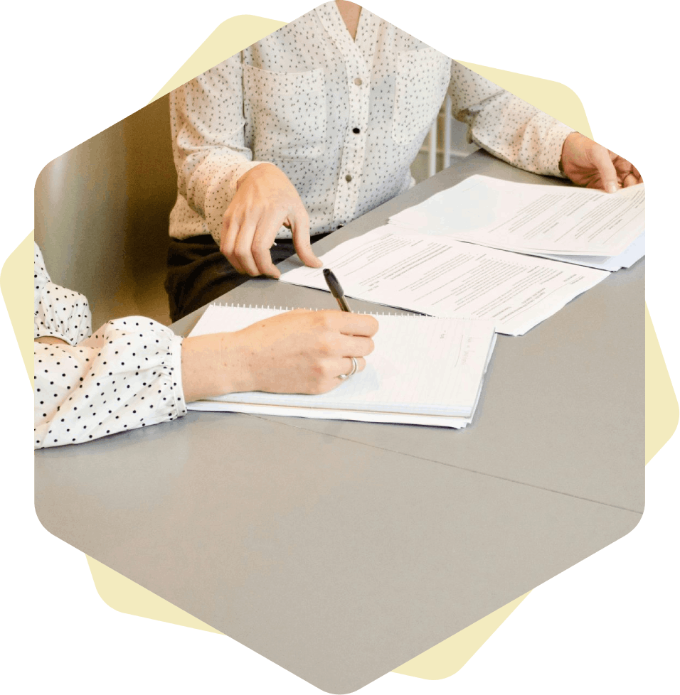 Mujer firmando en papel blanco de impresora junto a una mujer a punto de tocar los documentos.